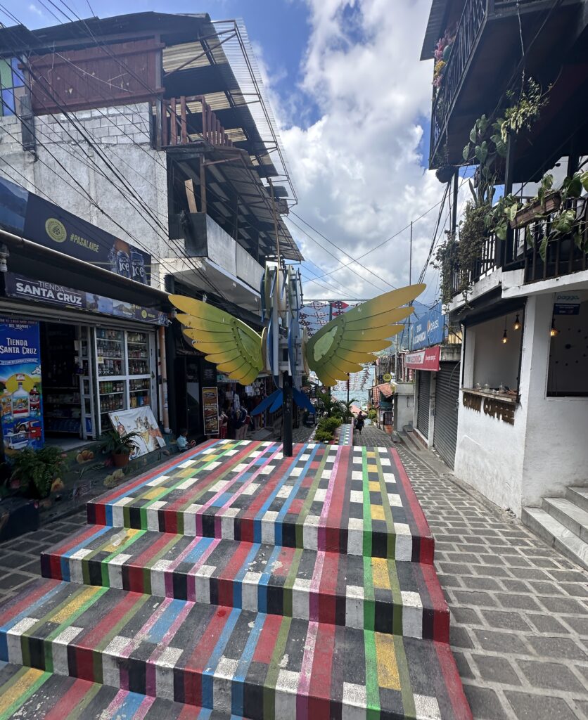 The image shows a colorful, striped staircase leading up to a platform with large, decorative butterfly wings. The wings are yellow and green, mounted on a pole. The scene is set in a narrow street lined with shops and buildings, some with balconies and plants. The sky is partly cloudy.
