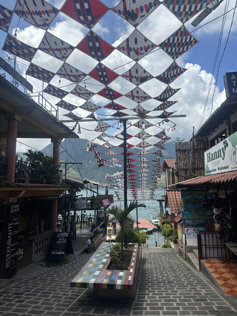 The image shows a narrow, cobblestone street in a small town, leading down to a body of water. Overhead, colorful, patterned fabric panels are strung across the street, creating a festive atmosphere. On either side of the street, there are buildings with signs, including a restaurant and a shop. A colorful bench with a small palm tree is in the center of the walkway. In the background, there are mountains and a partly cloudy sky.
