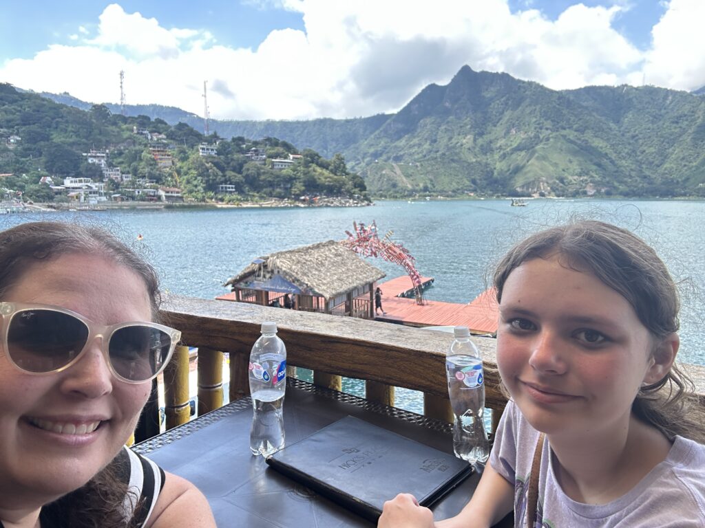 Two people are sitting at a table with bottled water in front of them, smiling at the camera. They are outdoors with a scenic view of a lake and mountains in the background. There are houses on the hillside and a thatched-roof structure near the water. The sky is partly cloudy.