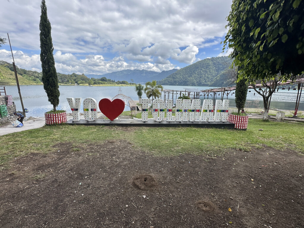 The image shows a scenic lakeside view with a large sign in the foreground that reads "YO ❤️ TOLIMÁN." The sign is colorful, with a red heart symbol in the middle. Behind the sign, there is a calm lake surrounded by lush green hills and a partly cloudy sky. There are trees and some structures near the water, adding to the picturesque setting.