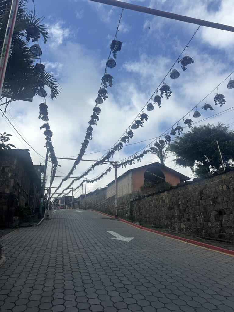 The image shows a paved street with a slight incline, lined with stone walls on both sides. Overhead, there are strings of decorative hanging ornaments, possibly lanterns or baskets, stretching across the street. The sky is partly cloudy, and there are some trees visible in the background.