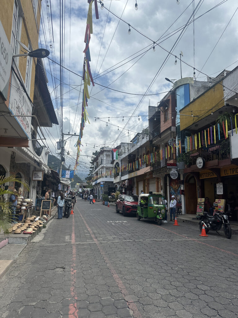 A lively street scene in a small town with cobblestone pavement. The street is lined with colorful flags and string lights overhead. On the left, there are shops displaying hats and other items, with people browsing. On the right, there are buildings with signs for a hotel and restaurant. A green tuk-tuk and a red car are parked on the street, and a few people are walking around. The sky is cloudy, adding a soft light to the scene.