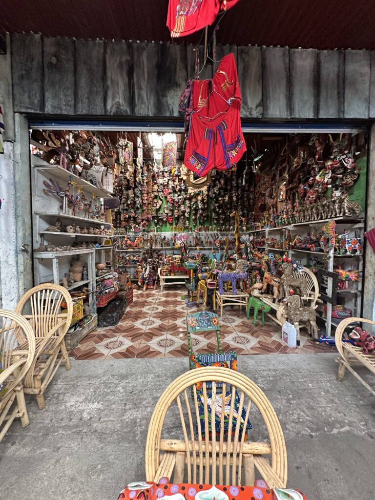 The image shows a colorful shop filled with various handcrafted items. The walls are lined with shelves displaying numerous masks, figurines, and pottery. Brightly painted wooden chairs and animal sculptures are arranged on the floor. Red garments hang from the ceiling, adding to the vibrant atmosphere. The shop has a tiled floor with a geometric pattern, and wicker chairs are placed at the entrance.
