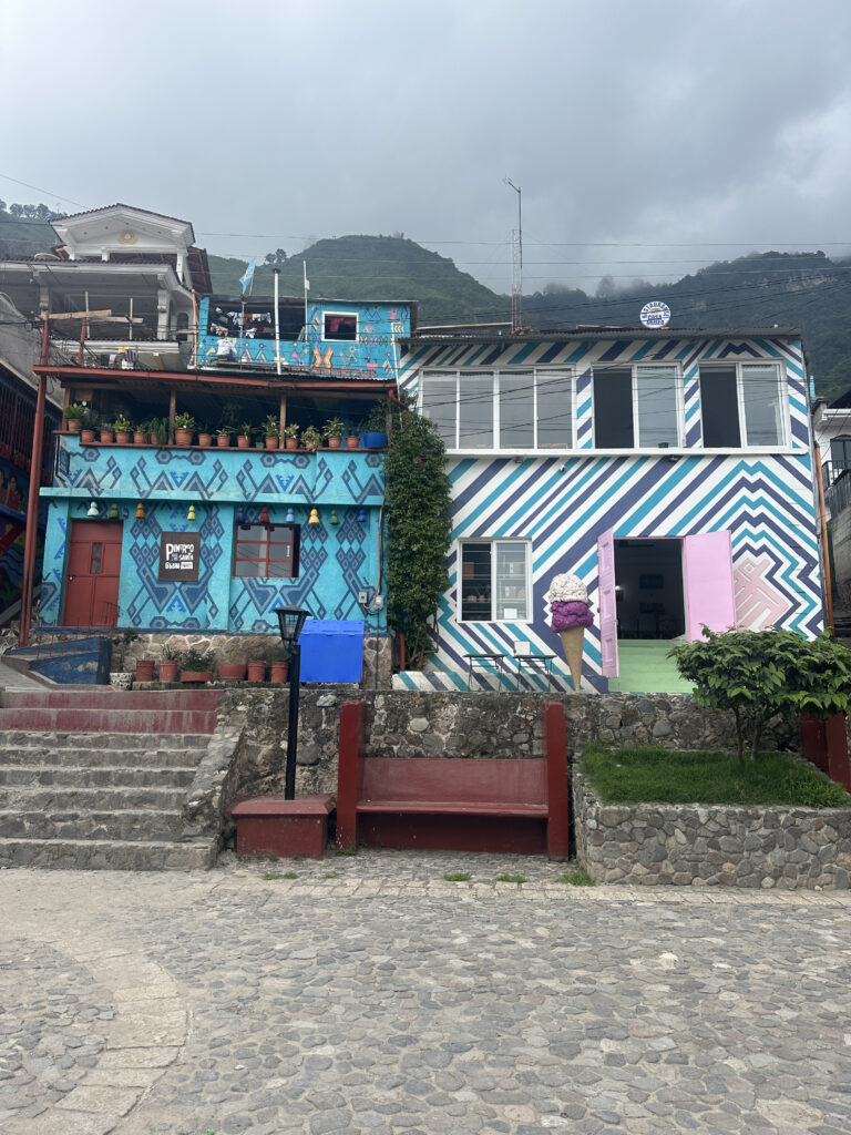 The image shows a colorful building with vibrant geometric patterns painted on its exterior walls. The left side of the building is decorated with blue and red designs, while the right side features blue and white diagonal stripes. There are potted plants on a balcony, and a mural of an ice cream cone is painted near a pink door. Stone steps and a cobblestone path lead up to the building, and there are red benches in front. In the background, there are green hills under a cloudy sky.