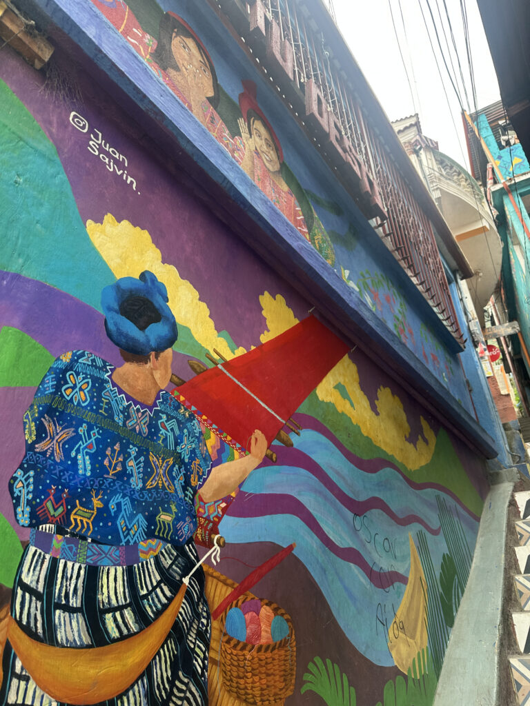 The image shows a vibrant mural painted on the side of a building. The mural features a person in traditional clothing, weaving with a red loom. The person is wearing a colorful, patterned garment and a blue headscarf. Below them is a basket filled with colorful yarn. The background includes abstract shapes and colors, such as purple, blue, and yellow, resembling a landscape. Above, there are two faces painted, and the name "Juan Sajvin" is signed on the mural. The scene is set in an urban environment with visible stairs and railings.