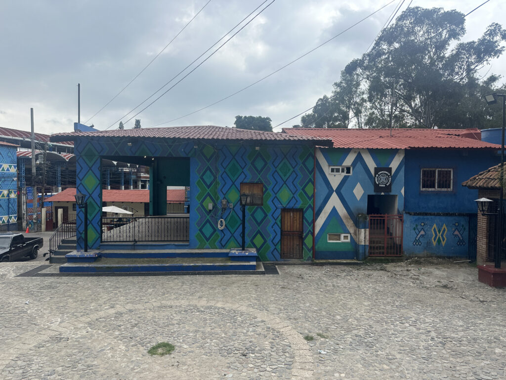 The image shows a colorful building with a vibrant geometric pattern painted on its exterior walls. The design features blue, green, and white diamond shapes and zigzags. The building has a red-tiled roof and a small entrance with a sign above it. The foreground consists of a cobblestone courtyard, and there are trees and a cloudy sky in the background.