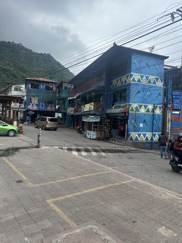 A street scene in a small town with colorful buildings. The building on the right is painted blue with geometric patterns and has a shop on the ground floor. There are several vehicles, including a green car and a motorcycle, parked on the street. People are walking and riding a scooter. In the background, there are lush green hills under a cloudy sky. Power lines are visible above the street.
