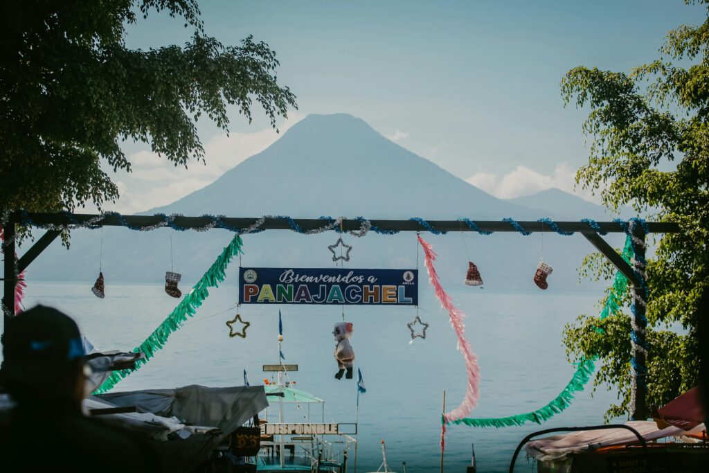 The image shows a scenic view of a lake with a large volcano in the background. In the foreground, there is a decorative archway with a sign that reads "Bienvenidos a Panajachel." The archway is adorned with colorful streamers, tinsel, and hanging decorations, including stars and small stockings. Trees frame the scene on both sides, and a boat is visible on the water below the sign.