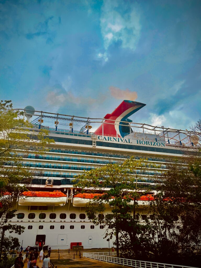 The image shows a large cruise ship named "Carnival Horizon" docked at a port. The ship has multiple decks with rows of windows and balconies. There are lifeboats visible on the side of the ship. The sky above is cloudy, and there are trees and people walking in the foreground.