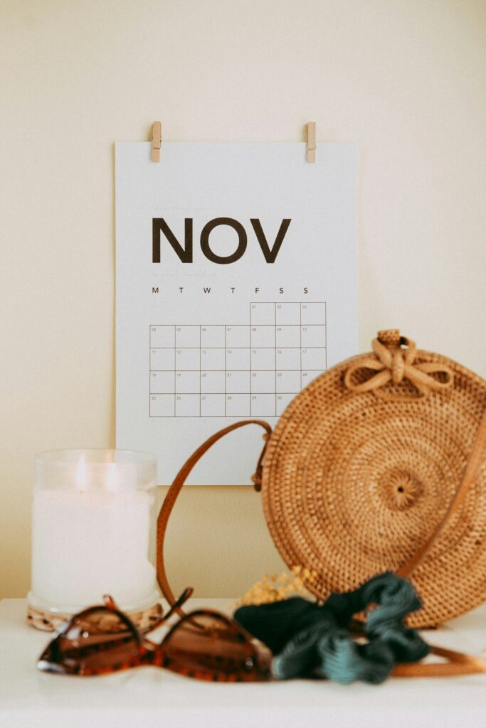 The image shows a November calendar page hanging on a wall. Below it, there is a white candle in a glass holder, a pair of tortoiseshell sunglasses, a woven round bag, and some fabric or ribbon. The setting has a warm, cozy aesthetic.