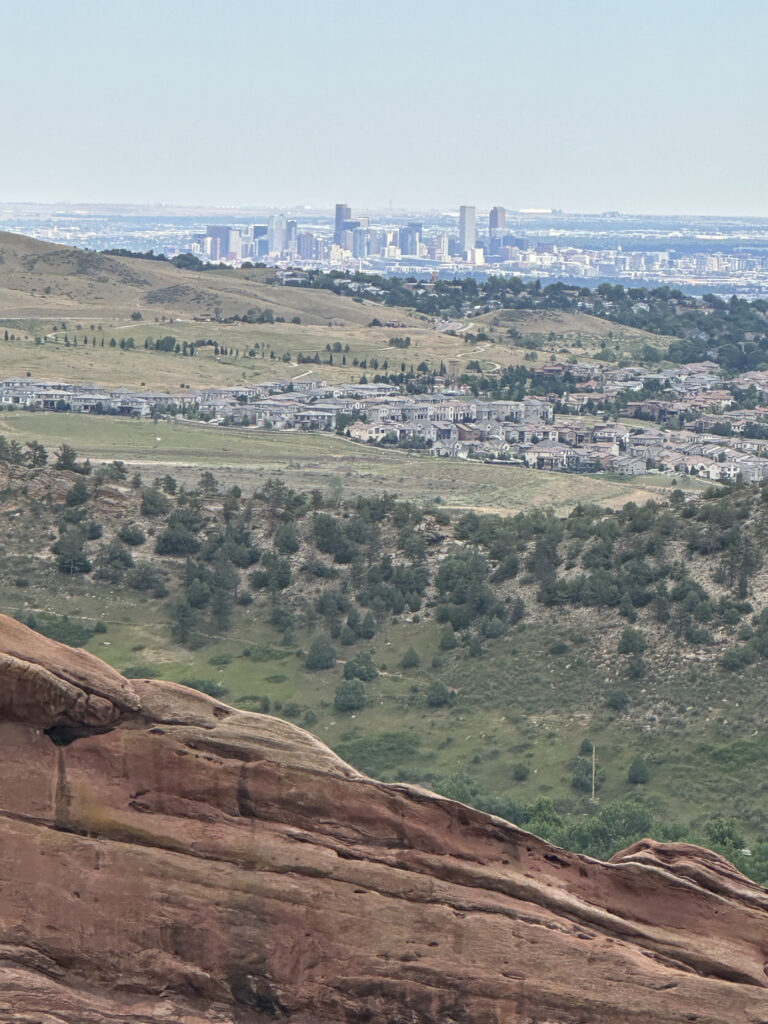 A scenic view of a city skyline in the distance, with a foreground of rolling hills and a large red rock formation. The landscape includes scattered trees and residential areas, with the city buildings visible under a clear sky.
