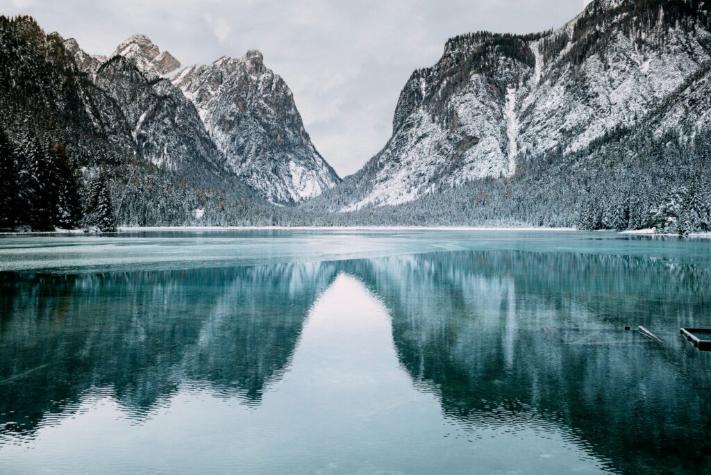 A serene winter landscape featuring a calm lake with a clear reflection of snow-covered mountains and pine trees. The mountains rise majestically in the background, and the scene is enveloped in a tranquil, overcast sky.