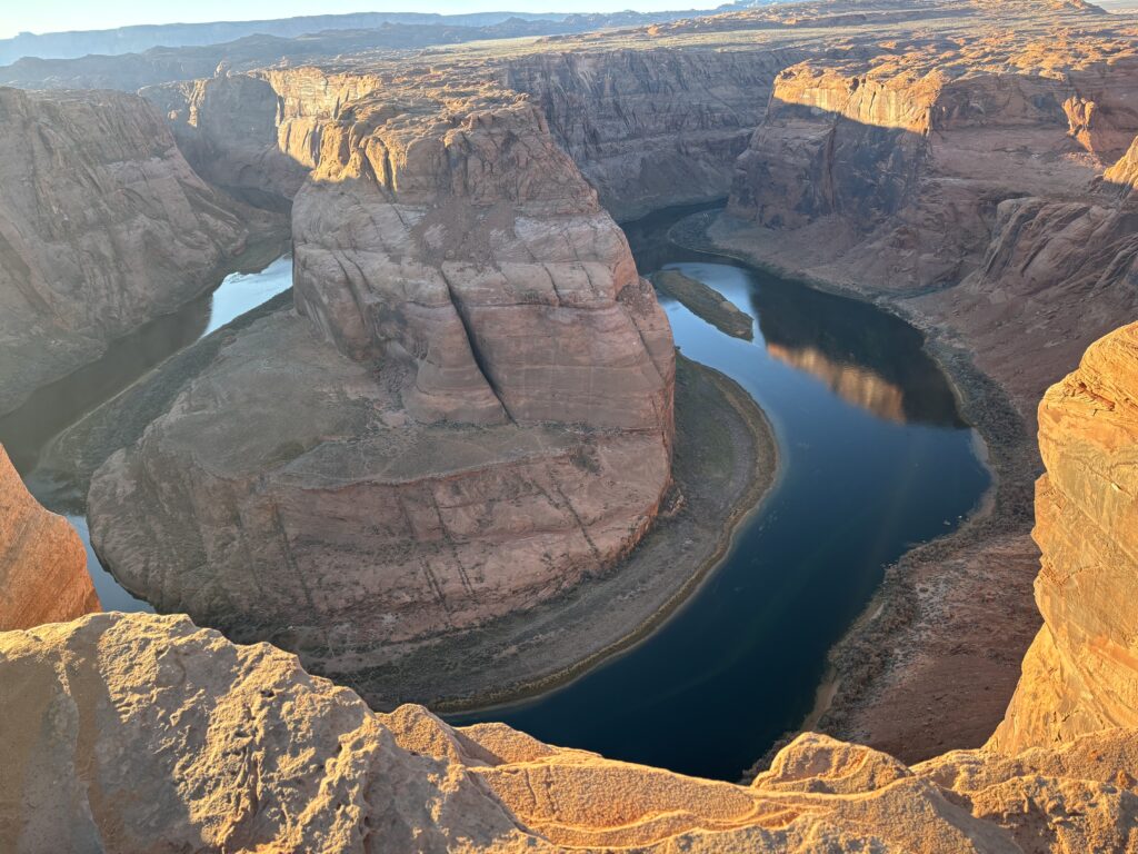 The image shows a scenic view of Horseshoe Bend, a famous meander in the Colorado River. The river curves around a large rock formation, creating a horseshoe shape. The surrounding landscape is composed of steep, reddish-brown cliffs and a clear blue sky is visible above. The lighting suggests it is either early morning or late afternoon, casting warm tones on the rocks.