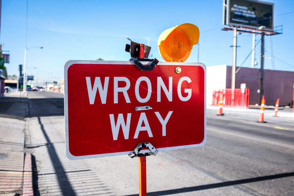 A red "WRONG WAY" sign is prominently displayed in the foreground, with an orange warning light attached to the top. The background shows a street with traffic cones and a billboard, under a clear blue sky.