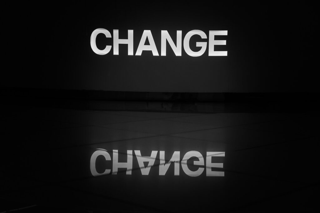 The image shows the word "CHANGE" in bold, white letters against a dark background. The word is reflected on a shiny surface below, creating a mirrored effect.