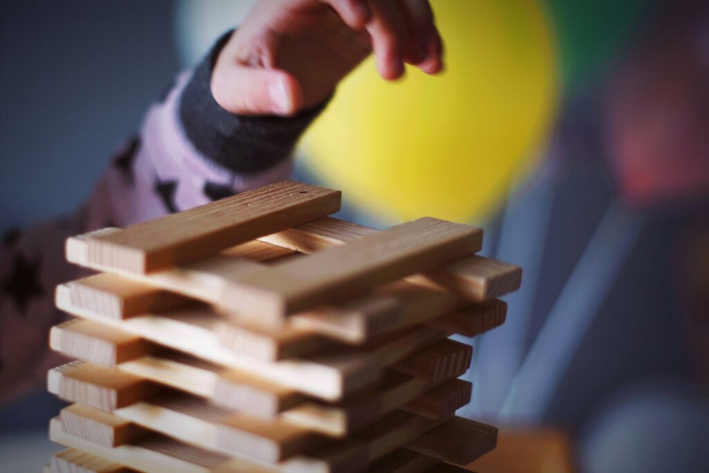 A child's hand is reaching towards a stack of wooden blocks arranged in a tower. The background is blurred with colorful shapes, including a yellow circle. The child is wearing a long-sleeved garment with a star pattern.