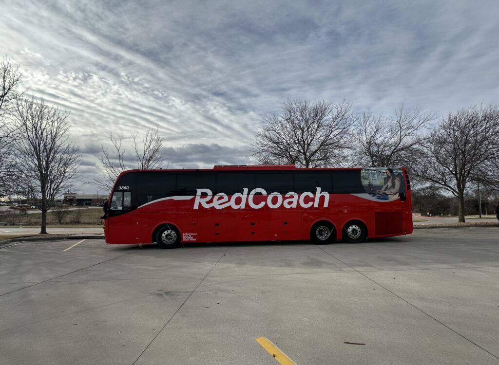 A large red bus with the word "RedCoach" written on its side is parked in an empty parking lot. The sky is overcast with streaks of clouds, and there are bare trees in the background. A person is standing near a white car on the right side of the image.