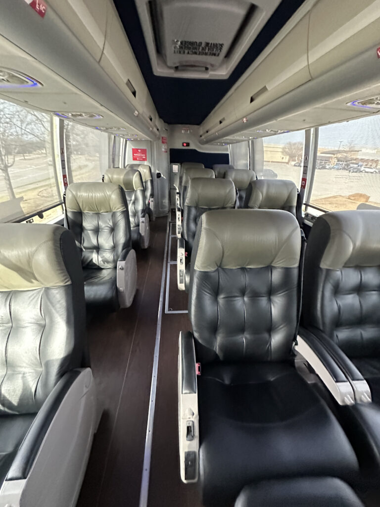 The image shows the interior of a bus with two rows of plush, leather seats. The seats are arranged in pairs on either side of a central aisle. The bus has large windows on both sides, allowing natural light to enter. The ceiling has overhead compartments and air vents. The overall appearance is clean and modern.
