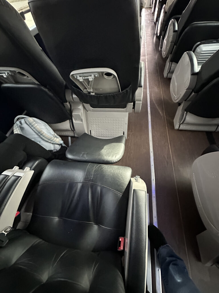 The image shows the interior of a bus or coach, featuring rows of black leather seats with armrests. The aisle runs down the middle, and a person's foot wearing a sock and jeans is visible in the aisle. A backpack is partially visible on the seat to the left.