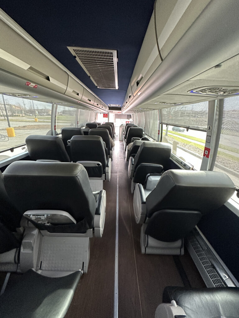 The image shows the interior of an empty bus. There are two rows of black leather seats on either side of a central aisle. The seats have armrests and are arranged to face forward. Overhead compartments are visible above the seats, and large windows line the sides of the bus, allowing natural light to enter. The floor appears to be made of wood or a wood-like material.