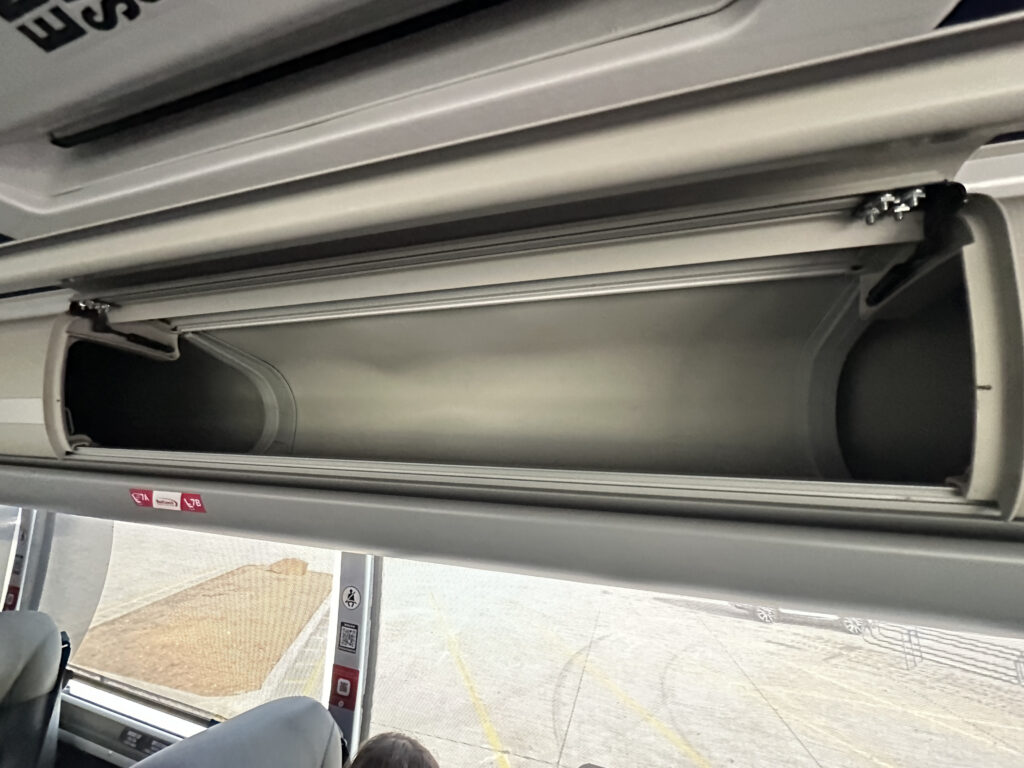 The image shows an empty overhead luggage compartment inside a bus. The compartment is open, revealing its interior. Below the compartment, part of a bus seat and a window are visible, with a view of the outside pavement.
