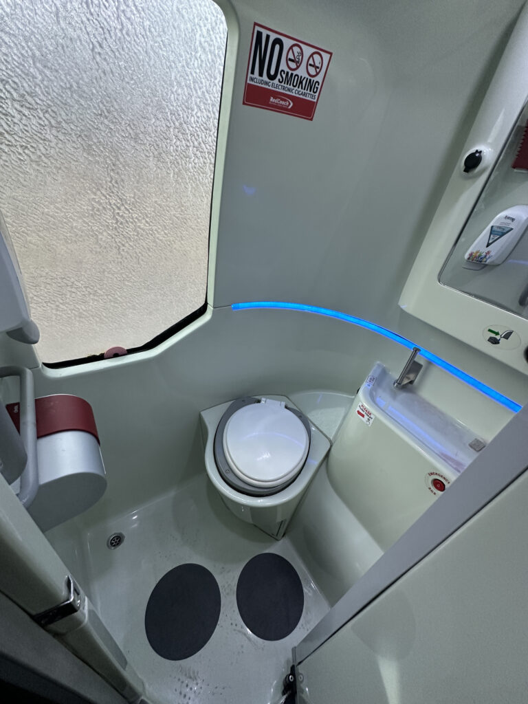 The image shows the interior of a compact airplane lavatory. It features a toilet with a closed lid, a small sink with a blue light above it, and a "No Smoking" sign on the wall. The floor has two black circular anti-slip mats, and there is a frosted window on the left side.