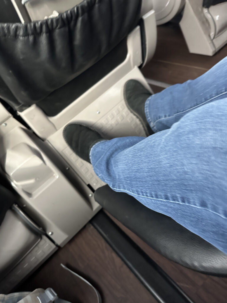 A person wearing blue jeans and black shoes is resting their feet on a footrest in a vehicle, possibly a bus or train. The interior features black and gray seating with a wooden floor.
