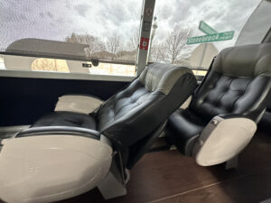 The image shows two black leather reclining seats inside a vehicle, possibly a bus or a train. The seats have white armrests and are positioned next to a large window. Outside the window, a street sign is visible, displaying the names "Stonebrook" and "Park." The background shows a cloudy sky and some trees.
