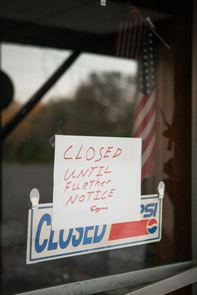 The image shows a glass door with two signs. The top sign is a handwritten note that reads "CLOSED UNTIL FURTHER NOTICE." Below it is a printed "CLOSED" sign with a Pepsi logo. An American flag is visible in the background.