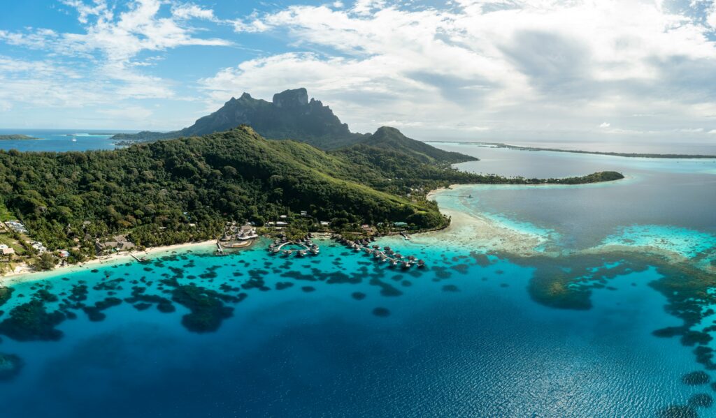 Aerial view of a tropical island with lush green mountains and a coastline surrounded by clear turquoise waters. Overwater bungalows are visible near the shore, and the ocean is dotted with coral reefs. The sky is partly cloudy, adding to the scenic beauty of the landscape.