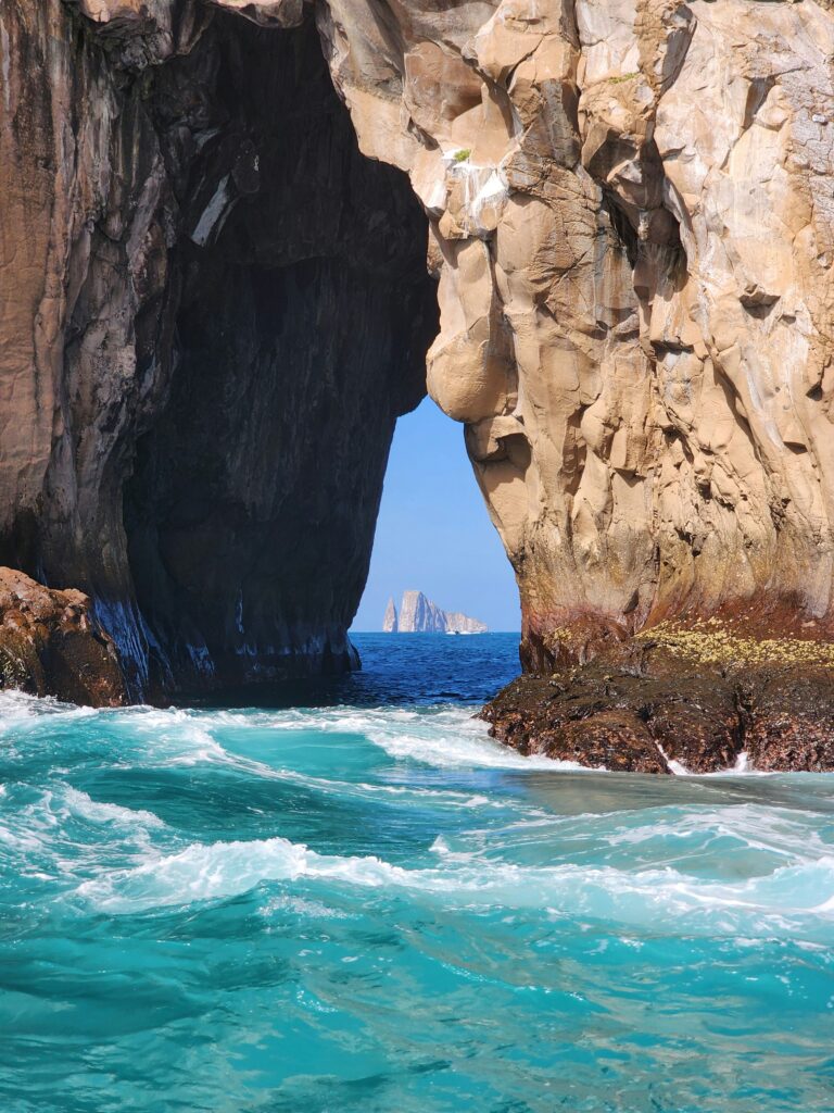 A natural rock archway over turquoise ocean water, with waves crashing against the rocks. Through the arch, a distant rock formation is visible against a clear blue sky.