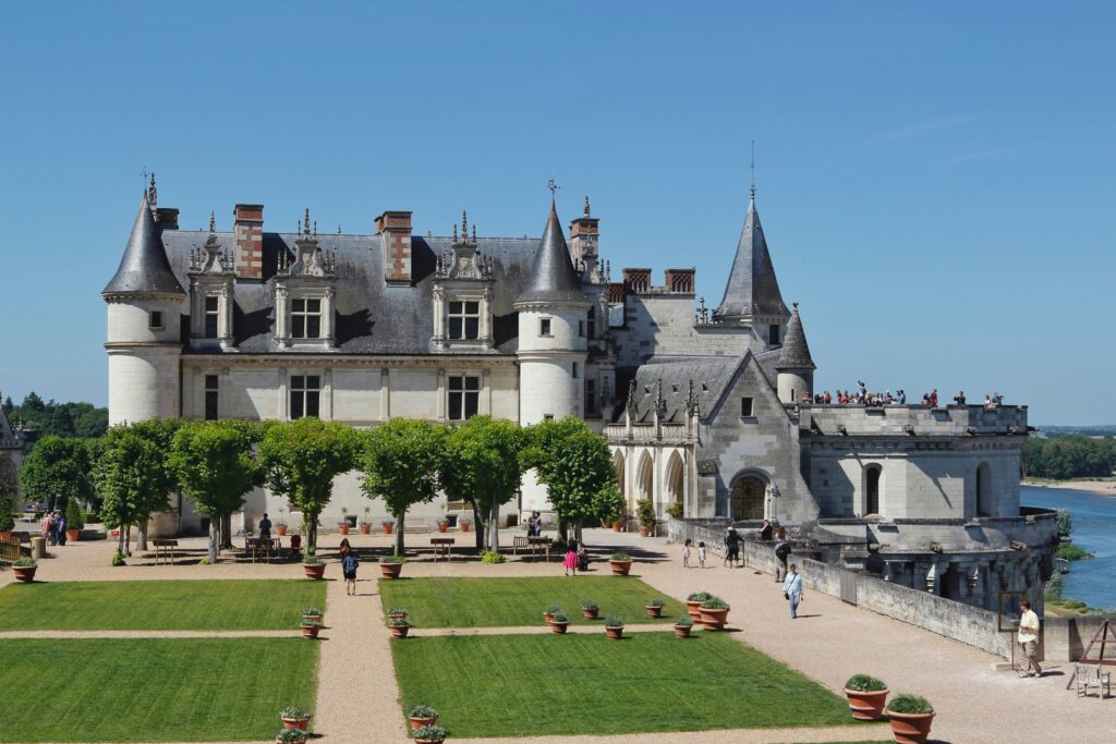 A historic castle with pointed towers and ornate architecture is set against a clear blue sky. The foreground features a well-maintained garden with neatly trimmed grass, potted plants, and a gravel pathway. Several people are walking and enjoying the scenery, and a group of visitors is gathered on a balcony overlooking a river.